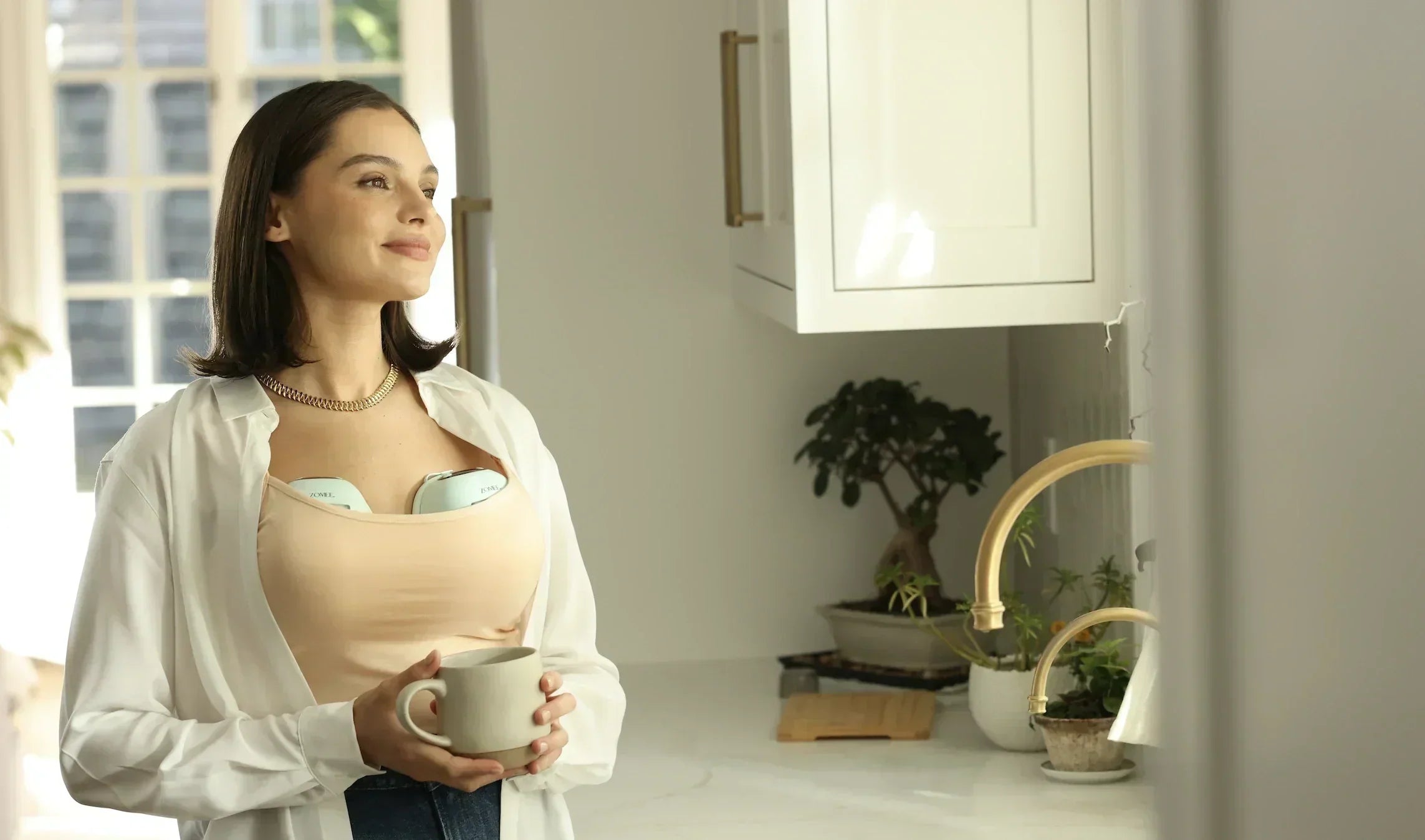 Mom wearing Zomee warming breast pump while holding a mug and looking out the window
