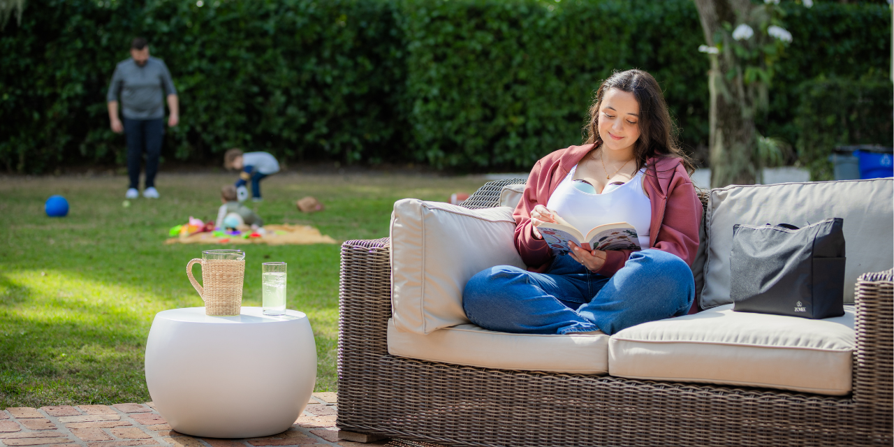 Woman reading on outdoor couch while wearing wearable breast pump, with family playing in background